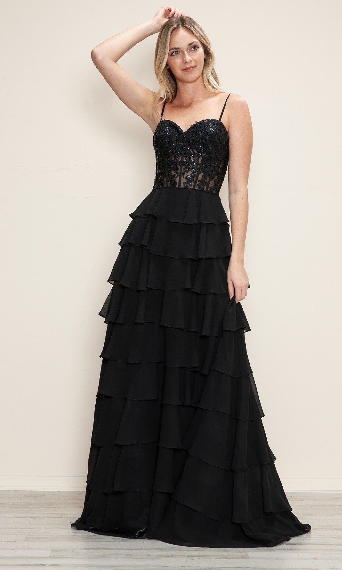 A woman wears the Poly USA 9698 Long Tiered Corset Prom Dress, featuring a sleeveless sequined bodice and a flowing tiered ruffle skirt, standing against a plain light-colored wall.